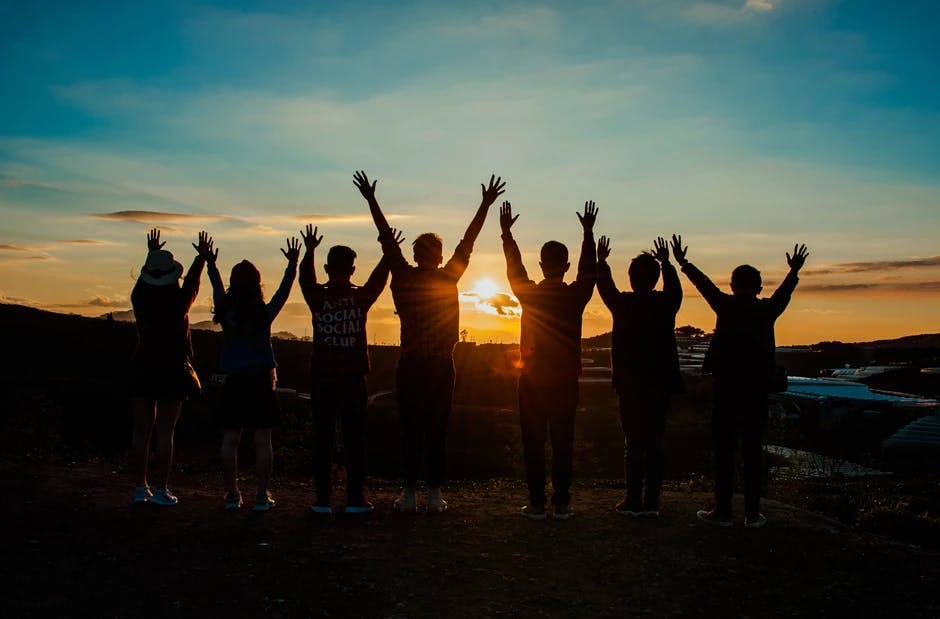 Image by Phan Minh. 7 Menschen, die vor einem Sonnenuntergang stehen und die Hände hochheben 